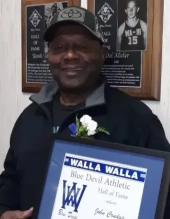 John Crudup, smiling in a cap with a white flower pinned to his jacket, holds a framed Walla Walla Blue Devil Athletic Hall of Fame certificate in front of hall of fame plaques.