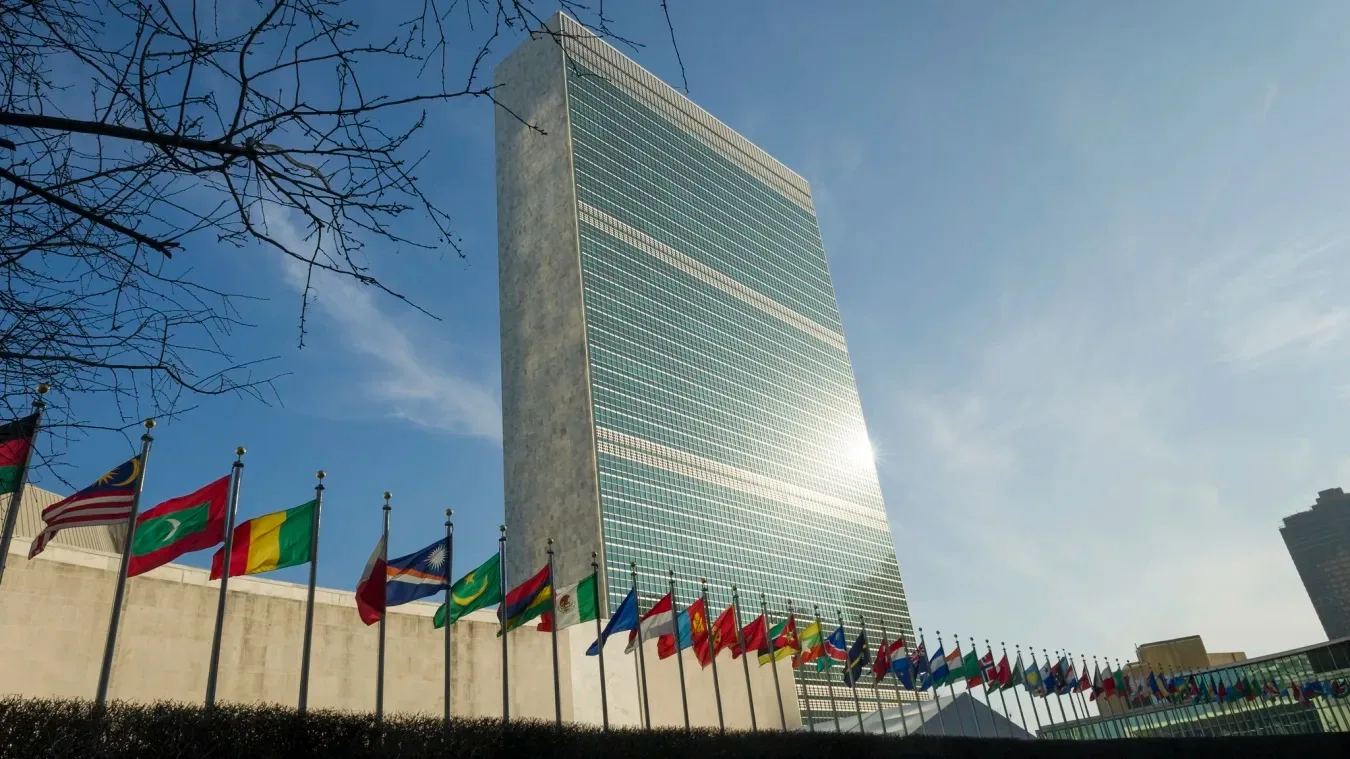 The tall glass-and-stone United Nations Secretariat building in New York under a blue sky, with a long row of national flags flying in front and bare tree branches at left.