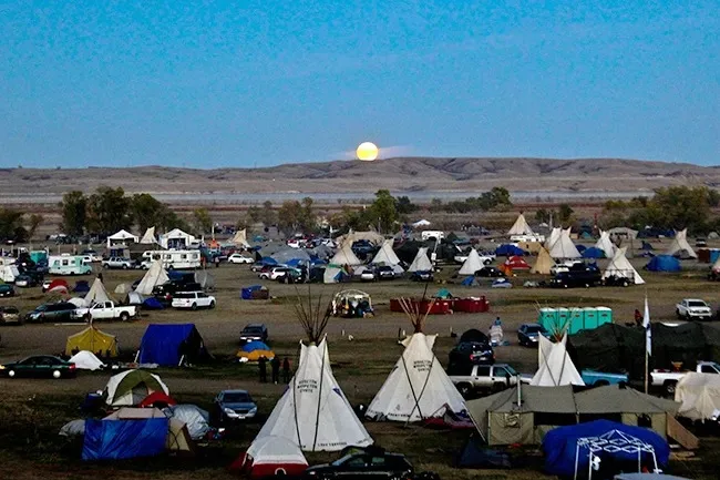 A full moon rises over hills above a large encampment of tipis, tents, vehicles and campers spread across a grassy plain at dusk.