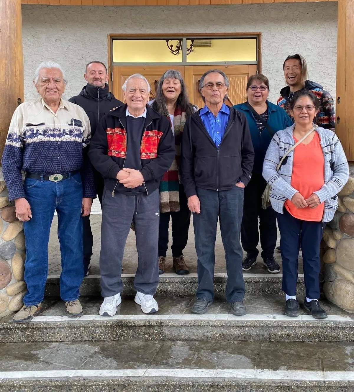 A group of eight smiling people, several of them elders, stand together on stone steps in front of the wooden double doors of a building.