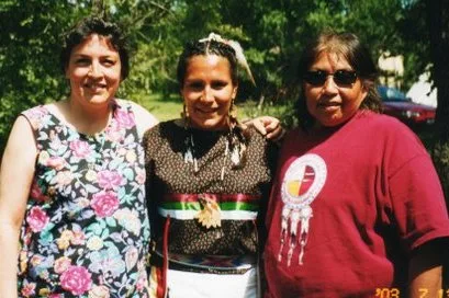 Three smiling women stand together outdoors among green trees: one in a floral dress, one in a beaded dress with a feather in her braided hair, and one in sunglasses and a red T-shirt.