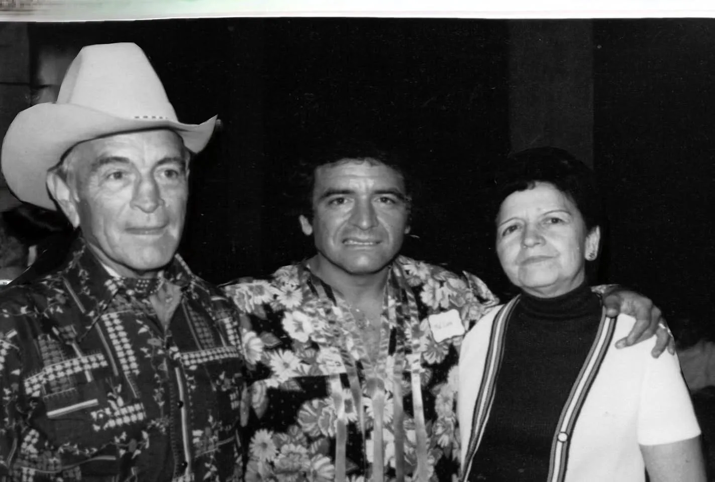 Black-and-white photograph of three people standing close: an older man in a white cowboy hat and patterned shirt, a younger man in a floral shirt with a name tag, and an older woman in a white cardigan.
