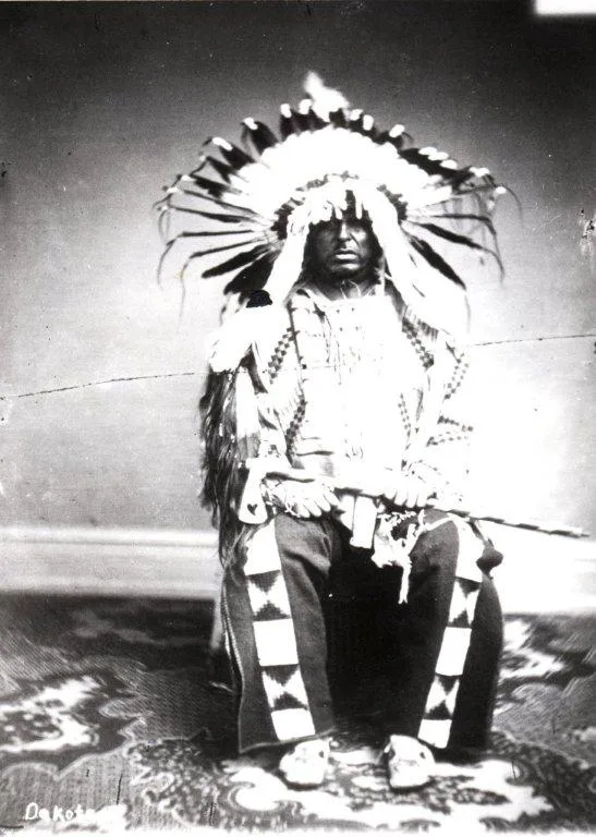 Black-and-white historical portrait of a Dakota man seated on a patterned rug, wearing a full eagle-feather headdress and beaded regalia, holding a long pipe across his lap.