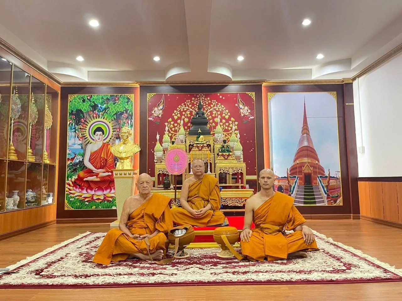 Three Buddhist monks in saffron robes sit on a patterned rug before a golden shrine with Buddha statues, flanked by murals of the Buddha and a red stupa.