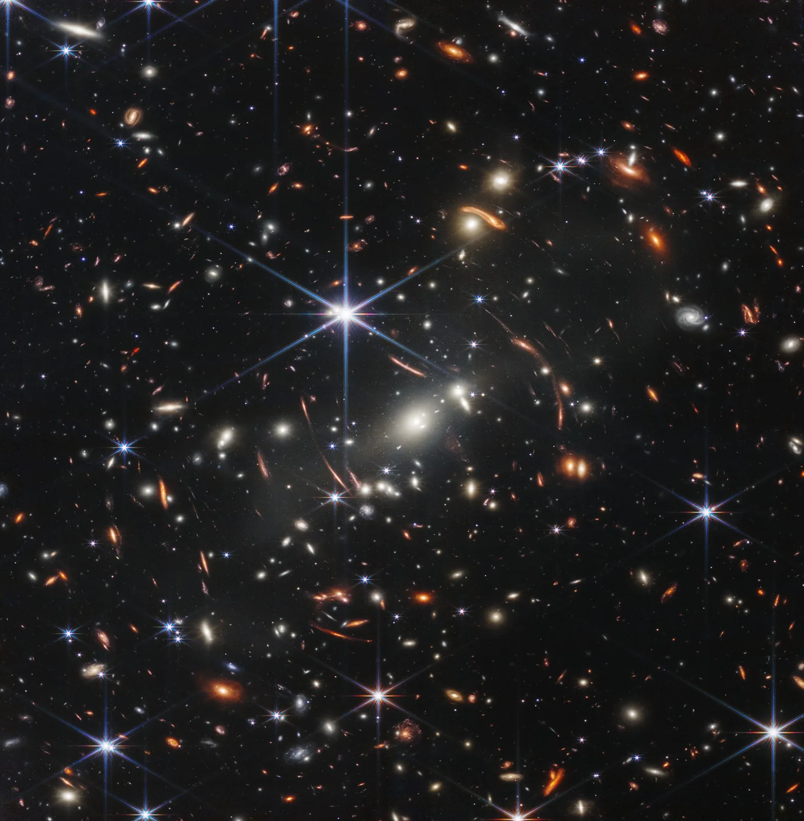 Deep field of space filled with countless glowing galaxies in white, gold and orange, some stretched into arcs, with bright stars showing six-pointed spikes against black sky.