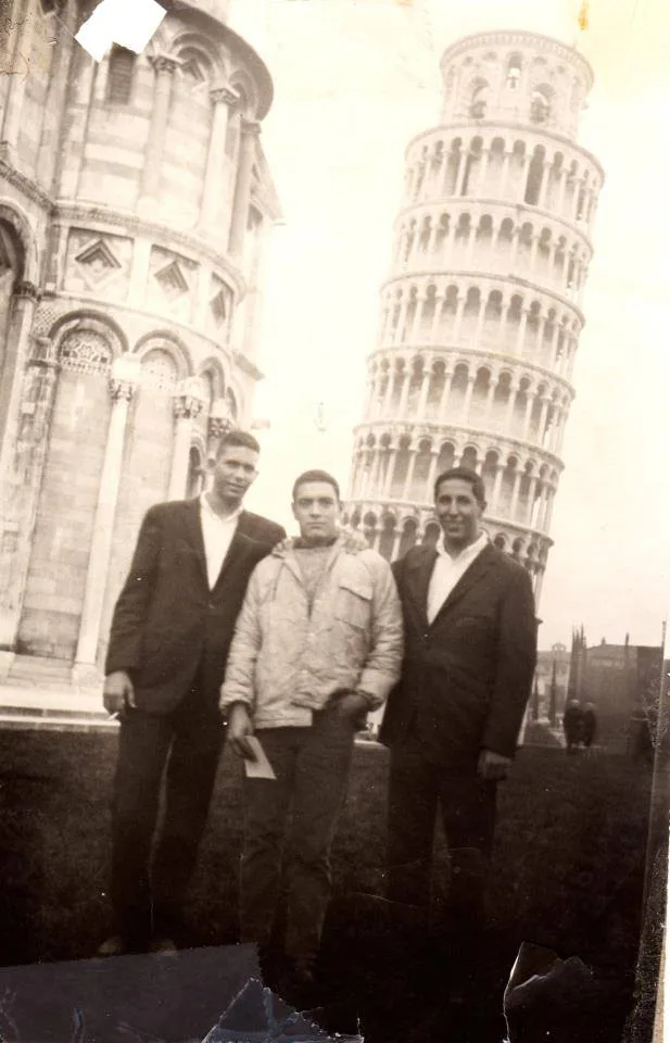 Old black-and-white photograph of three young men in jackets standing together in front of the Leaning Tower of Pisa.