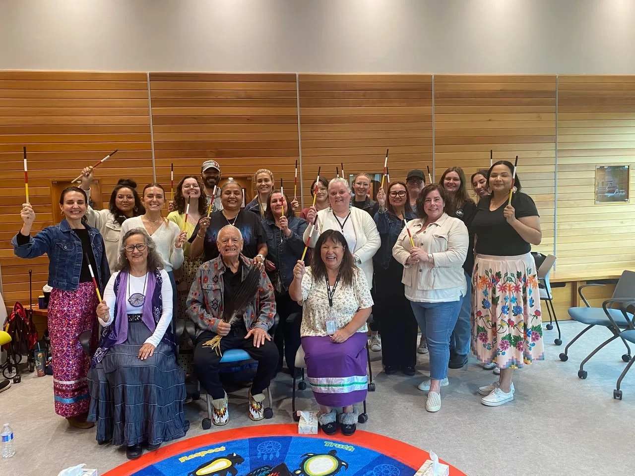 A group of about twenty people, mostly women, pose in a wood-paneled room, many raising red, yellow, black and white sticks. A seated elder holds an eagle feather fan.