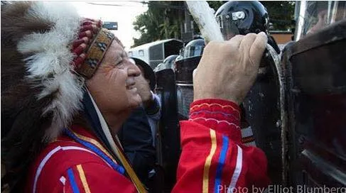 A smiling elder in a feather headdress and red ribbon shirt presses his hand against riot police shields in a street. Photo credit: Elliot Blumberg.