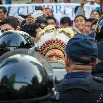 An elder in a beaded feather headdress with painted face stands among helmeted riot police, a crowd with phones and a banner behind him.
