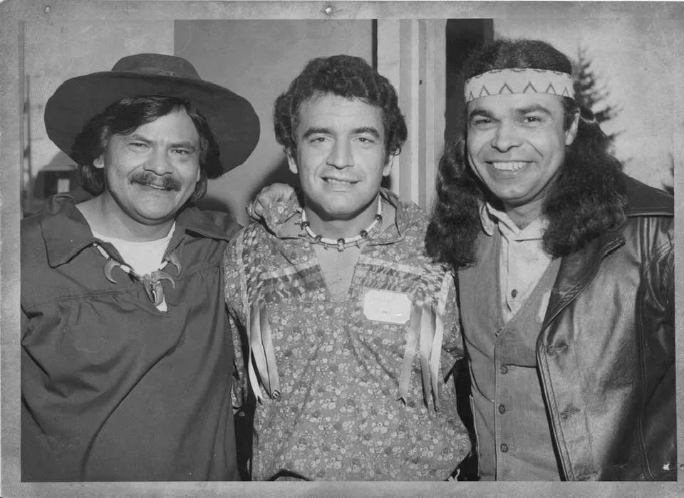 Black-and-white photo of three smiling men standing arm in arm: one in a wide-brimmed hat with a claw necklace, one in a floral ribbon shirt, and one with long hair and a beaded headband.