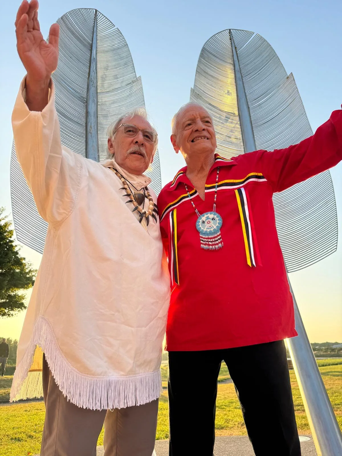 Phil Lane Jr. and Angaangaq, one in a red ribbon shirt and one in a fringed white tunic, raise their arms before tall steel feather sculptures at sunset.