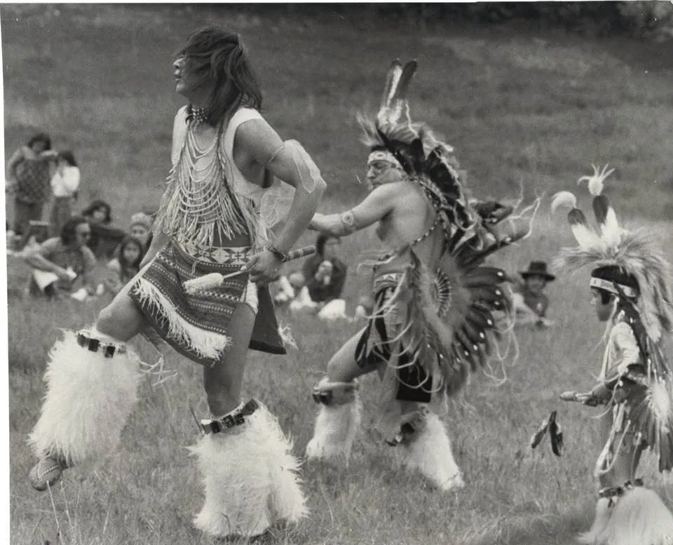 Black-and-white photo of three dancers in regalia with white fur leg bands dancing on a grassy field, one in a feather bustle and a child in feathers, as spectators sit on the grass behind.