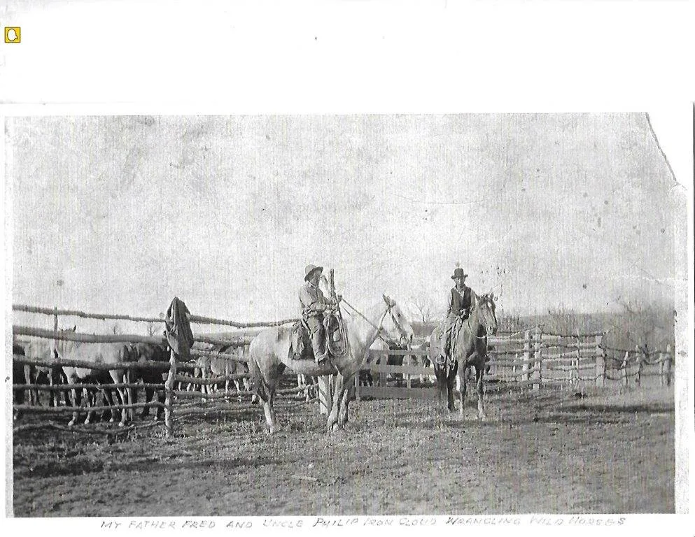 Old black-and-white photograph of two men on horseback in hats beside a wooden rail corral holding horses. A handwritten caption reads: My father Fred and Uncle Philip Iron Cloud wrangling wild horses.