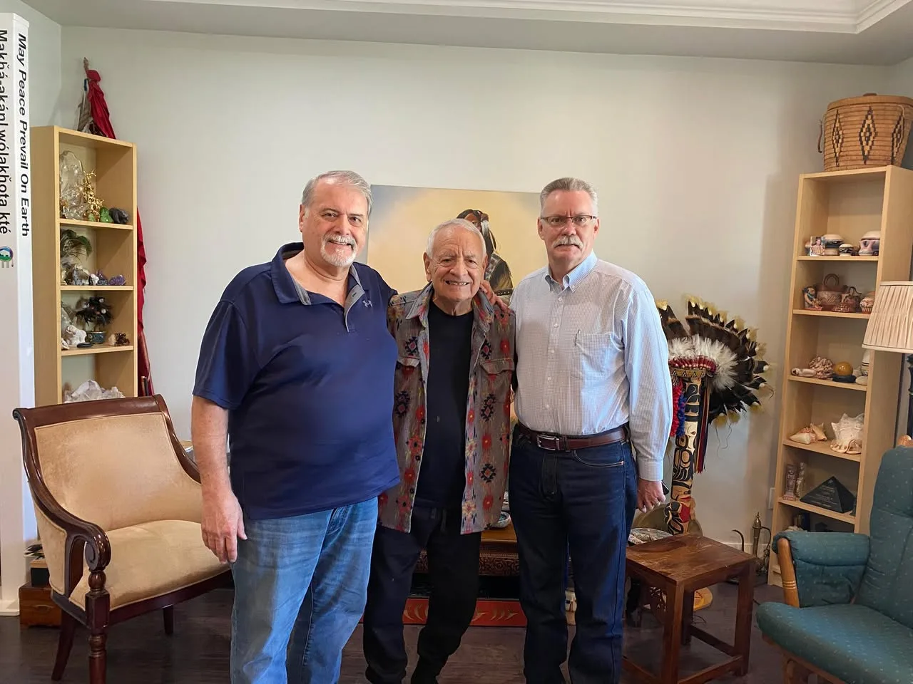 Three men stand together smiling in a living room: an elder in a patterned jacket between two men, with a feather headdress, carved pole, shelves of crafts and a 'May Peace Prevail On Earth' pole.