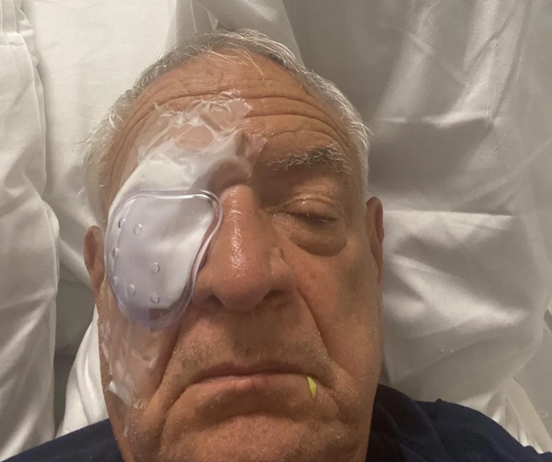 An older man with gray hair lies on a white hospital pillow, a clear protective shield taped over his right eye, his left eye closed.