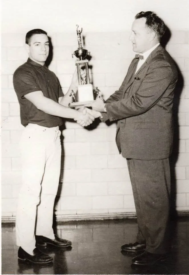 Black-and-white photo of a young Phil Lane Jr. in a dark polo shirt shaking hands with Coach McBride, in a suit and bow tie, as he presents a tall wrestling trophy in a gym.