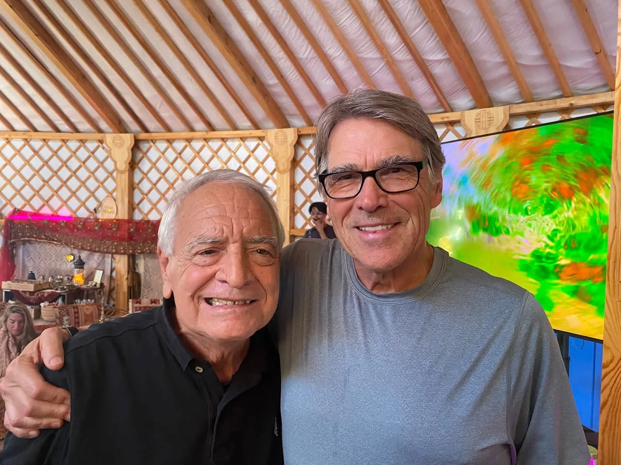 Two smiling older men stand arm in arm inside a yurt with a wooden lattice frame, one in a black shirt and one in glasses and a gray shirt, beside a colorful screen.