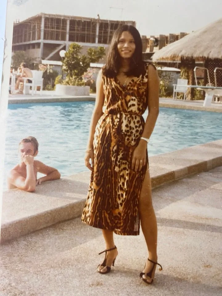 A vintage photograph of Suthida, a young woman with long dark hair in a leopard-print sleeveless dress and strappy heels, standing smiling beside a hotel swimming pool.