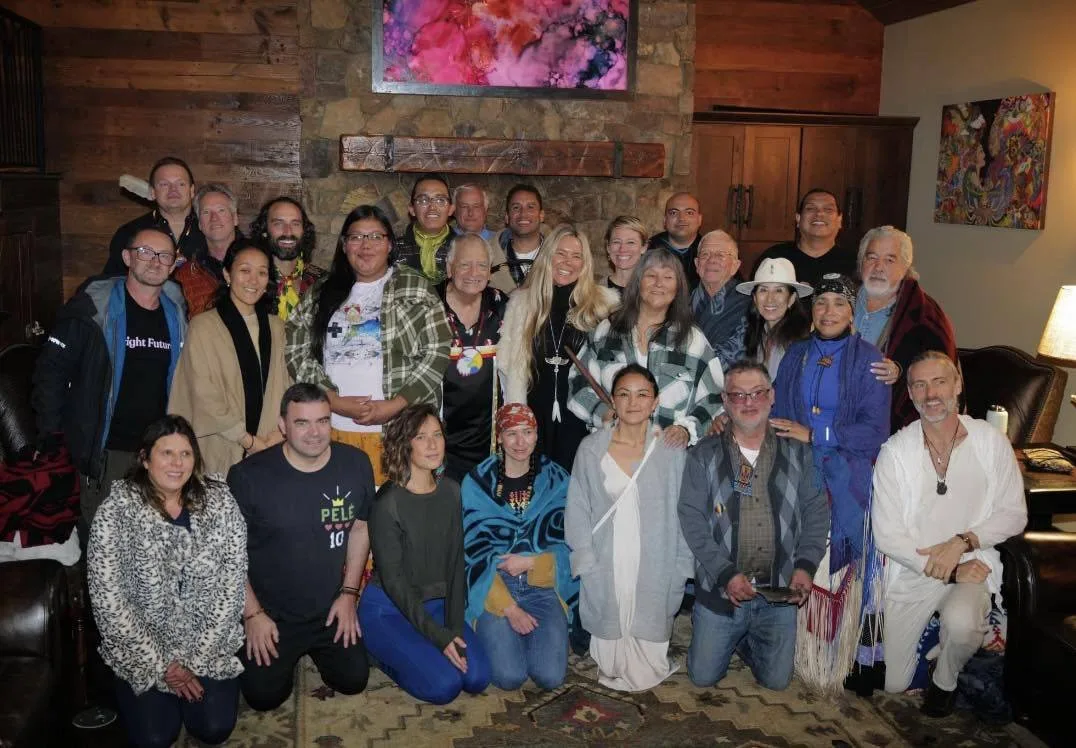 A large group of about thirty people, some in traditional Indigenous dress and blankets, gather for a portrait in a wood-paneled room with a stone fireplace and colorful paintings.