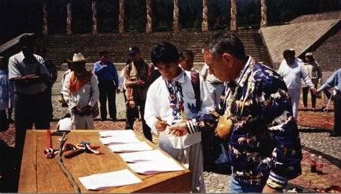 At a stone amphitheater, a man in a feathered shirt and bead necklace and a young man in an embroidered white shirt stand at a table with documents, watched by a gathered crowd.