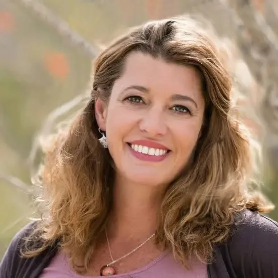 Portrait of a smiling woman with shoulder-length wavy light brown hair, wearing a pink top, pendant necklace and silver earrings, outdoors with a soft blurred background.