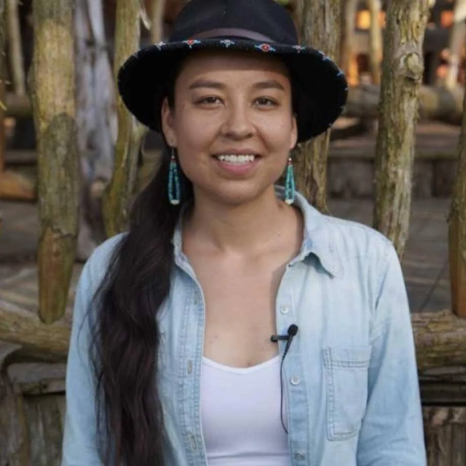 A smiling young Indigenous woman with a long dark braid, wearing a black beaded-brim hat, turquoise earrings and a denim shirt, seated before a rustic log structure.