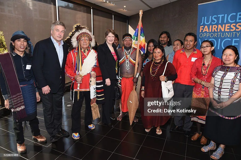 A group of Indigenous leaders and supporters in traditional dress stand together indoors, including an elder in a feather headdress holding a fan, a man holding a rainbow Wiphala flag on a paddle, and people in suits, beside a banner reading Justice, Fairness, Opportunity, Voice.