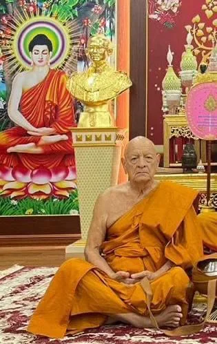 Phil Lane Jr., head shaved and wearing saffron monk's robes, sits cross-legged on a carpet before a golden bust and a painting of the Buddha on a lotus.