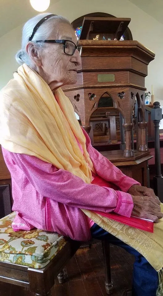 Hazel Red Bird, an elderly woman with white hair and glasses, wearing a pink top and pale yellow shawl, sits on a floral cushioned stool beside a carved wooden church pulpit.