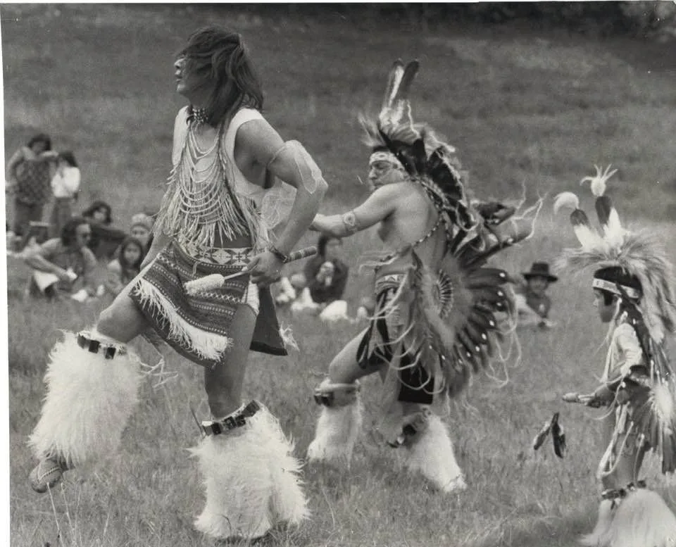 Black-and-white photo of dancers in regalia on a grassy field: one in a beaded cape and fur leggings, one in a feather bustle and headdress, and a young dancer, with onlookers seated behind.
