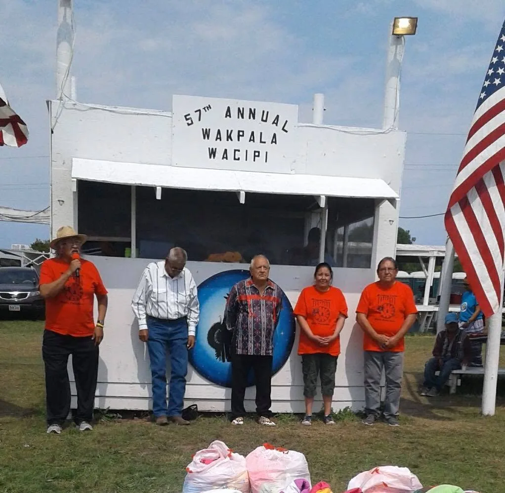 Five people stand on the grass before a white announcer's stand with a sign reading "57th Annual Wakpala Wacipi"; an elder in an orange shirt and straw hat speaks into a microphone beside an American flag.