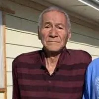 An elderly man with short gray hair, wearing a maroon striped shirt, stands outside in front of a light-colored house.