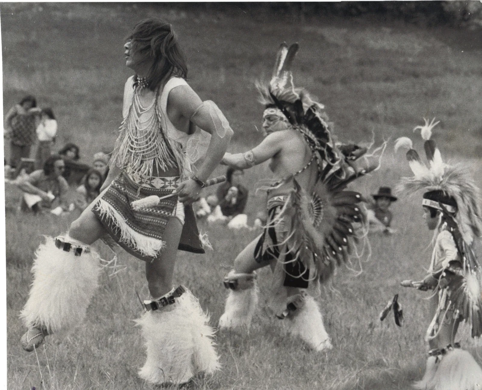 Black-and-white photo of dancers in regalia, with fur leggings, feathered bustles and headdresses, dancing on grass at Discovery Park as people sit watching. Phil and Sam are among them.