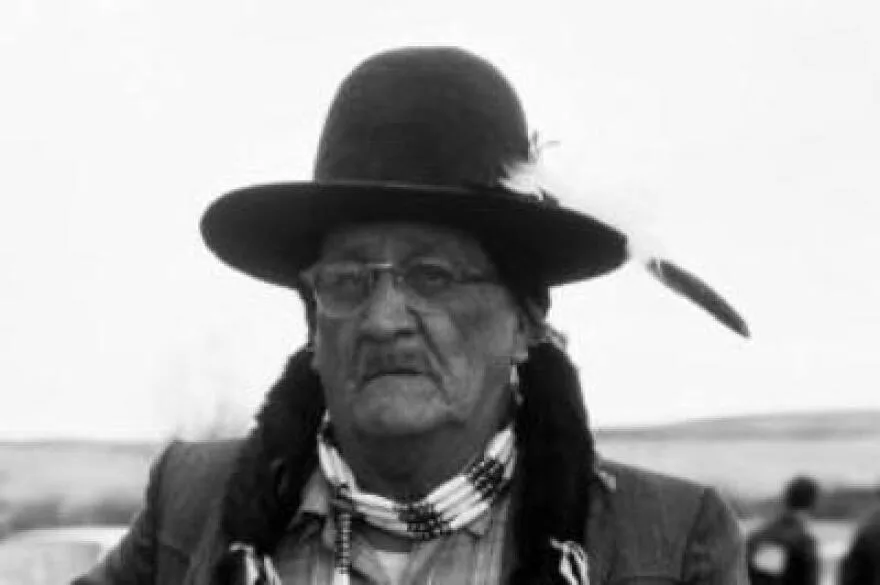 Black-and-white portrait of an elder wearing glasses, a black brimmed hat with a feather, a beaded choker and fur-wrapped braids, standing outdoors on open prairie.