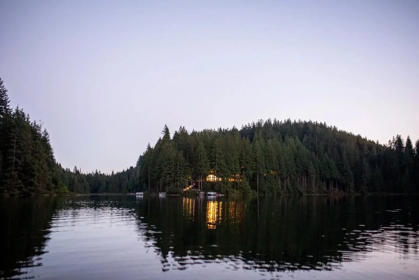 A calm lake at dusk surrounded by dark evergreen forest, with a lit lodge among the trees reflecting golden light on the water.