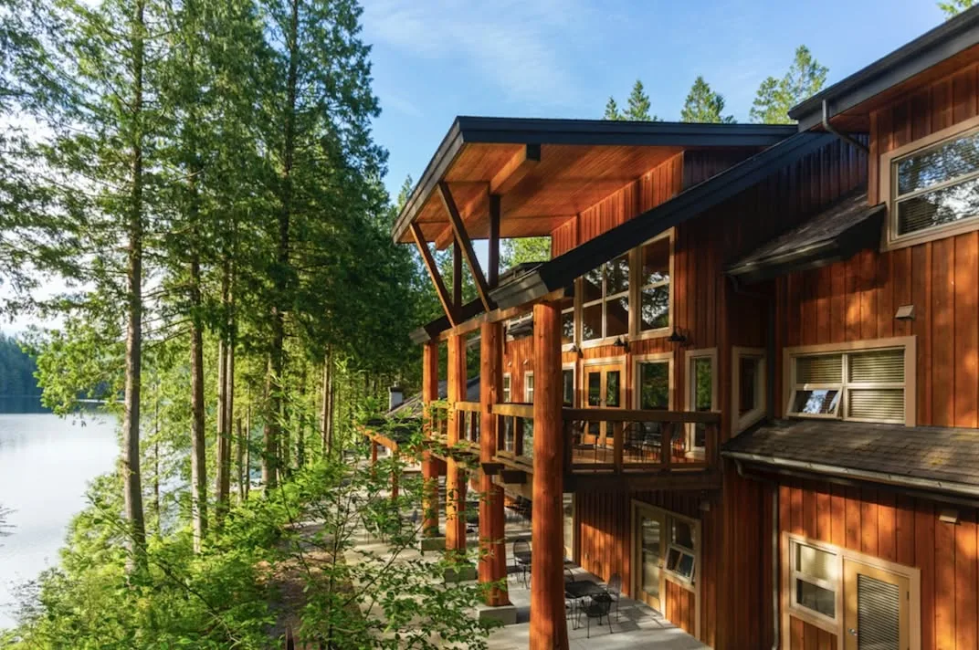 A large wooden lodge with timber posts and a covered deck, set among tall evergreens beside a lake on a sunny day.