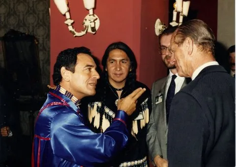 A man in a blue satin ribbon shirt gestures as he speaks to an older man in a dark suit at an indoor reception, while a man with long braids in a beaded, fringed shirt and others look on.