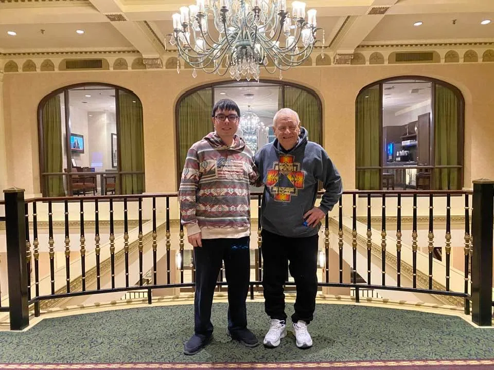 Phil Lane Jr. and his son Tiger stand together at a railing on a hotel mezzanine beneath a crystal chandelier, both wearing hoodies with Indigenous-style designs.