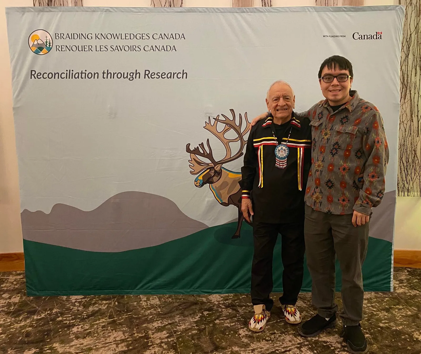 Phil Lane Jr., an elder in a black ribbon shirt, beaded medallion and moccasins, stands with his son Tiger before a Braiding Knowledges Canada banner reading "Reconciliation through Research."