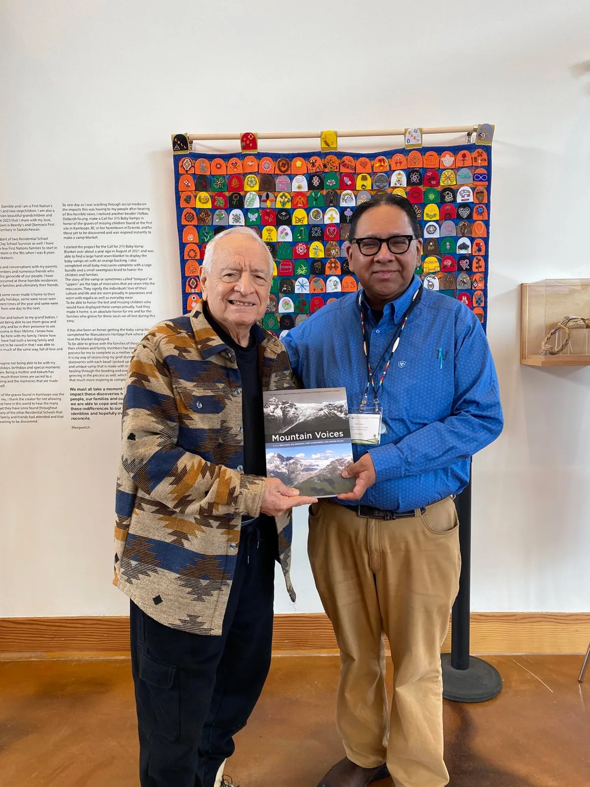 Two smiling men hold a copy of the book Mountain Voices in front of a large blue blanket covered in colourful beaded baby moccasin vamps.