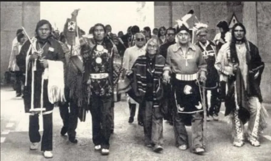Black-and-white photo of Indigenous representatives in traditional regalia, some holding staffs and feathers, walking together with a crowd behind them.