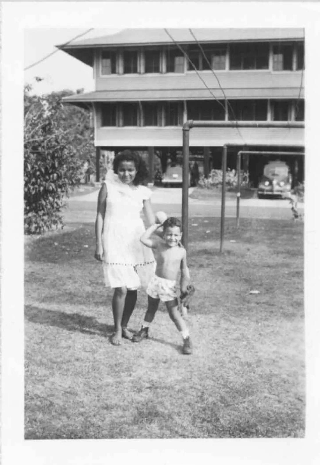 Black-and-white photo of Dominga, a young woman in a white dress, standing on a lawn beside a smiling small boy in shorts, with a raised two-story house and vintage cars behind them.