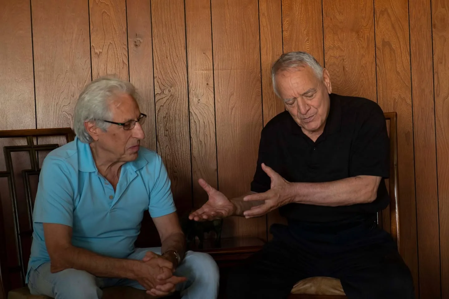 Two older men seated against a wood-paneled wall in conversation; the man in a black shirt gestures with both hands as the man in a blue shirt listens.