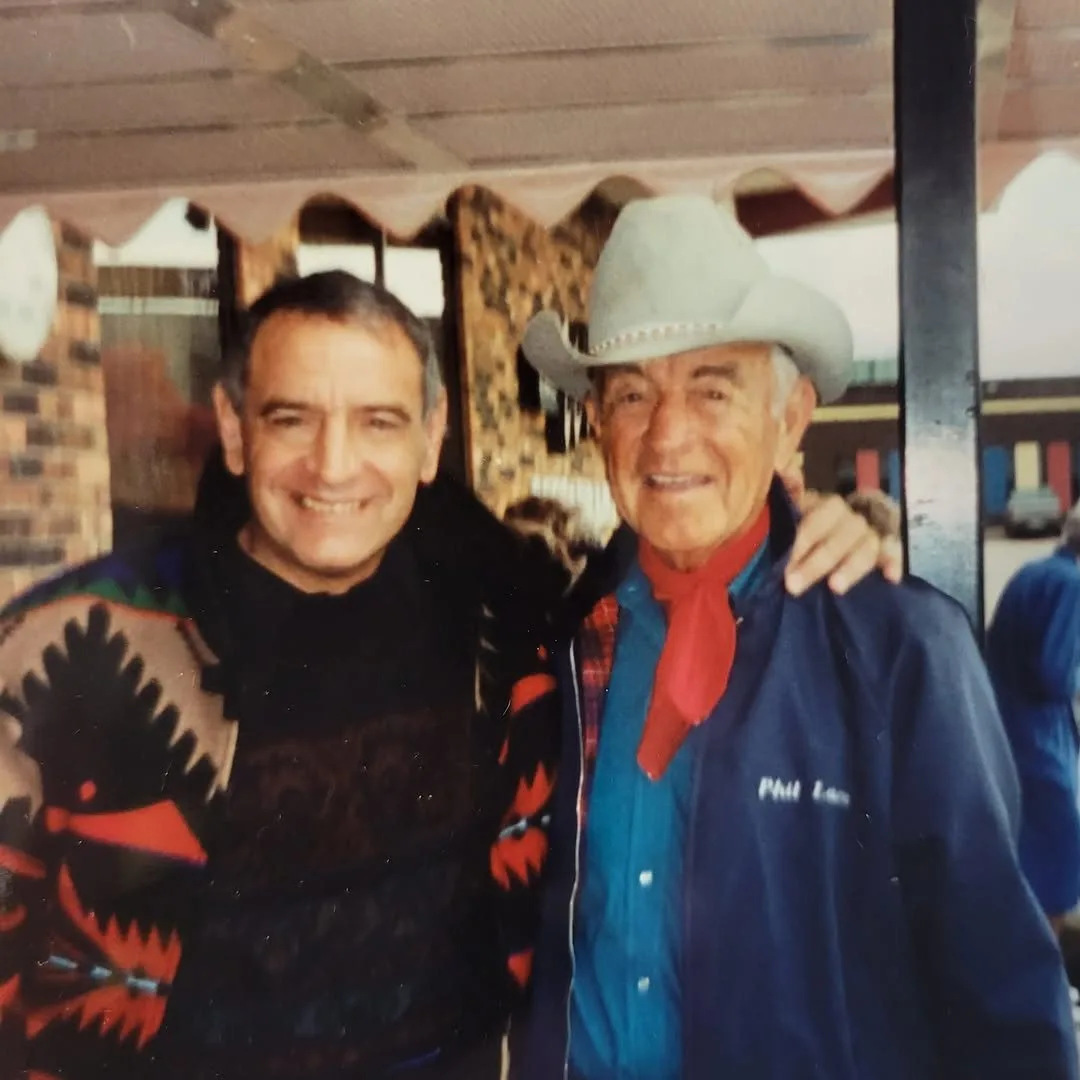 Phil Lane Jr., smiling in a patterned sweater, with his arm around his father, who wears a light cowboy hat, red neckerchief and a navy jacket.