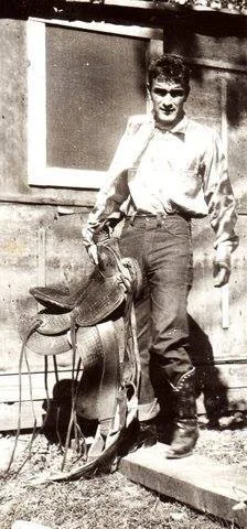 Vintage black-and-white photo of a young man in a light shirt, jeans and boots, carrying a saddle beside a wooden building.