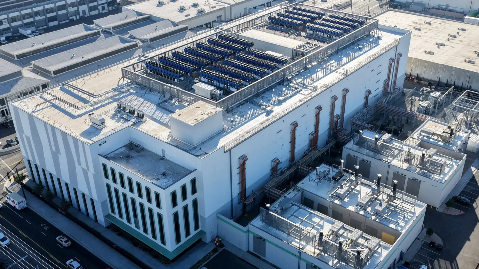 Aerial view of a large white data center building with rows of blue cooling units on its roof, surrounded by generator buildings, electrical equipment and city streets.
