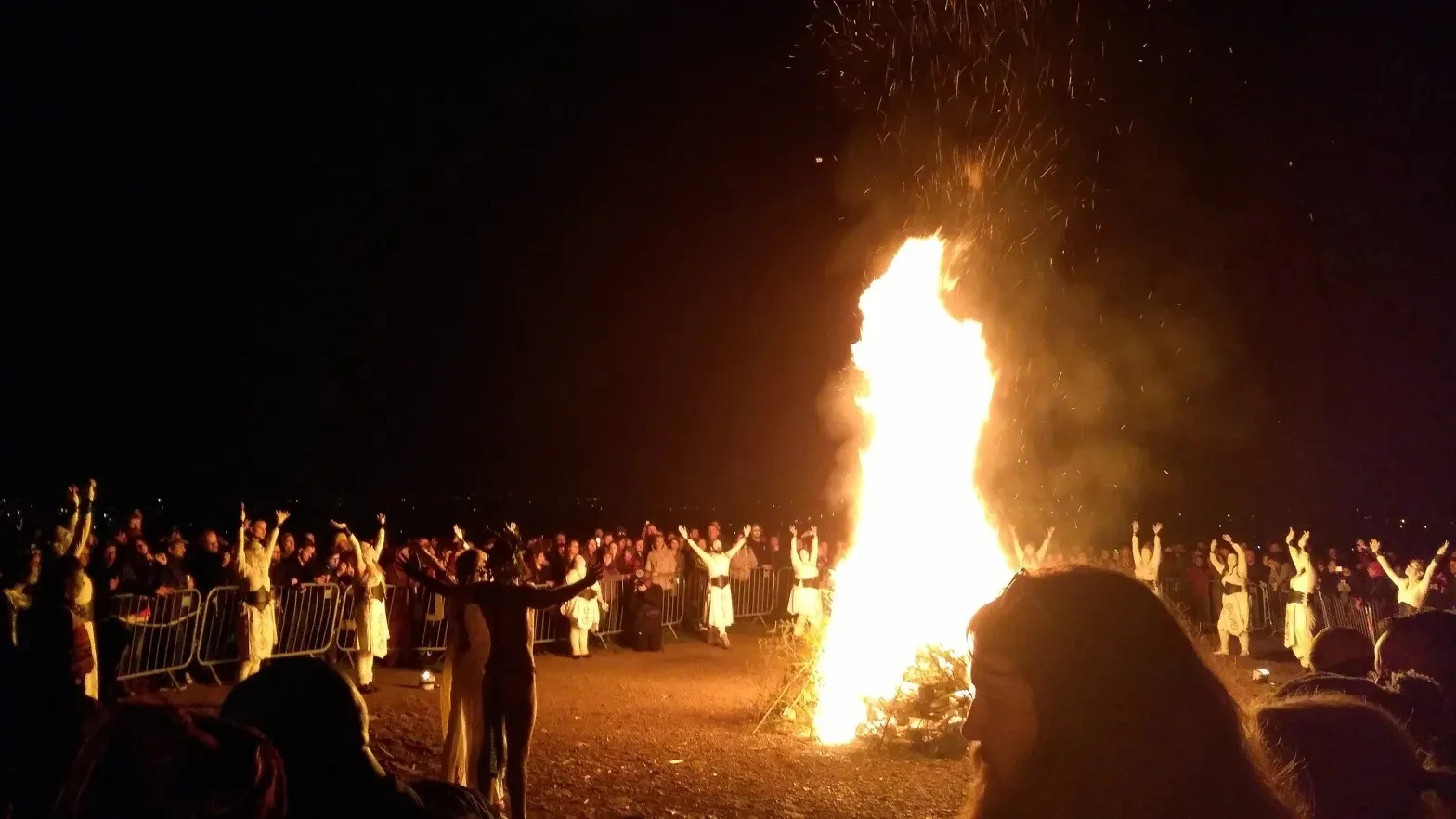 A towering bonfire blazes at night, sending sparks skyward, as white-robed performers with raised arms circle it before a large crowd behind barriers.
