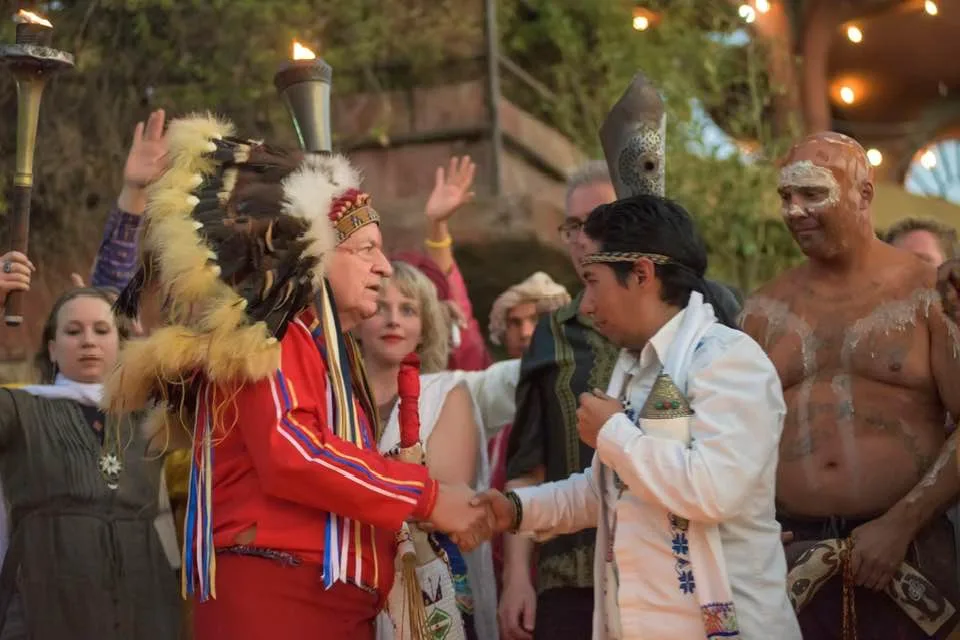 Phil Lane Jr., in a feather headdress and red regalia, shakes hands with Otomi Brother Mindahi in a white embroidered shirt, among a crowd holding torches, with Jason Kelly in body paint at right.