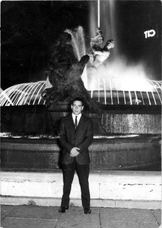 Black-and-white night photo of a young man in a dark suit and tie standing before a lit fountain with bronze sculptures in Rome.
