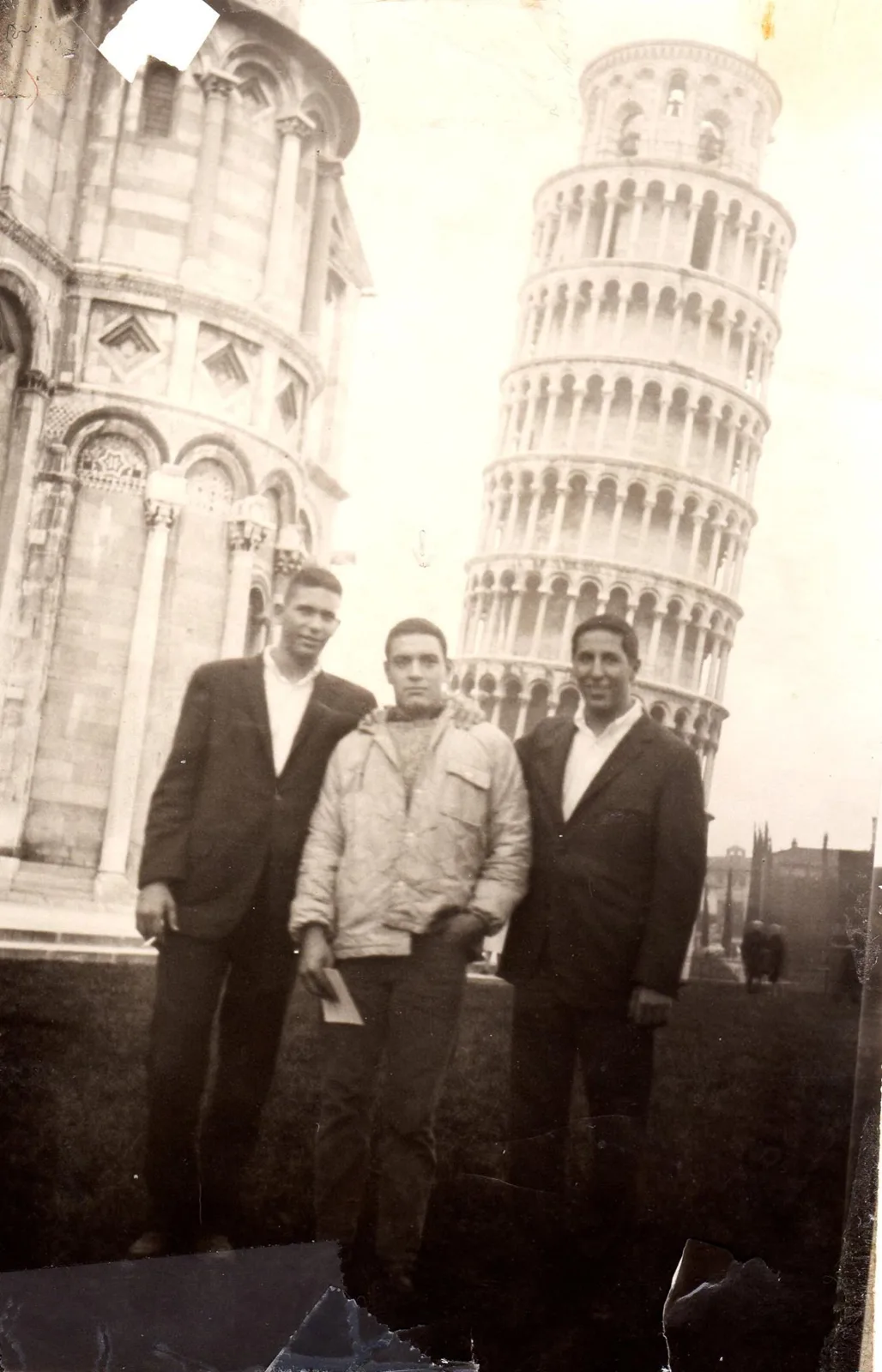 Faded black-and-white photo of three young men standing together on a lawn, with the Leaning Tower of Pisa and a marble building behind them.